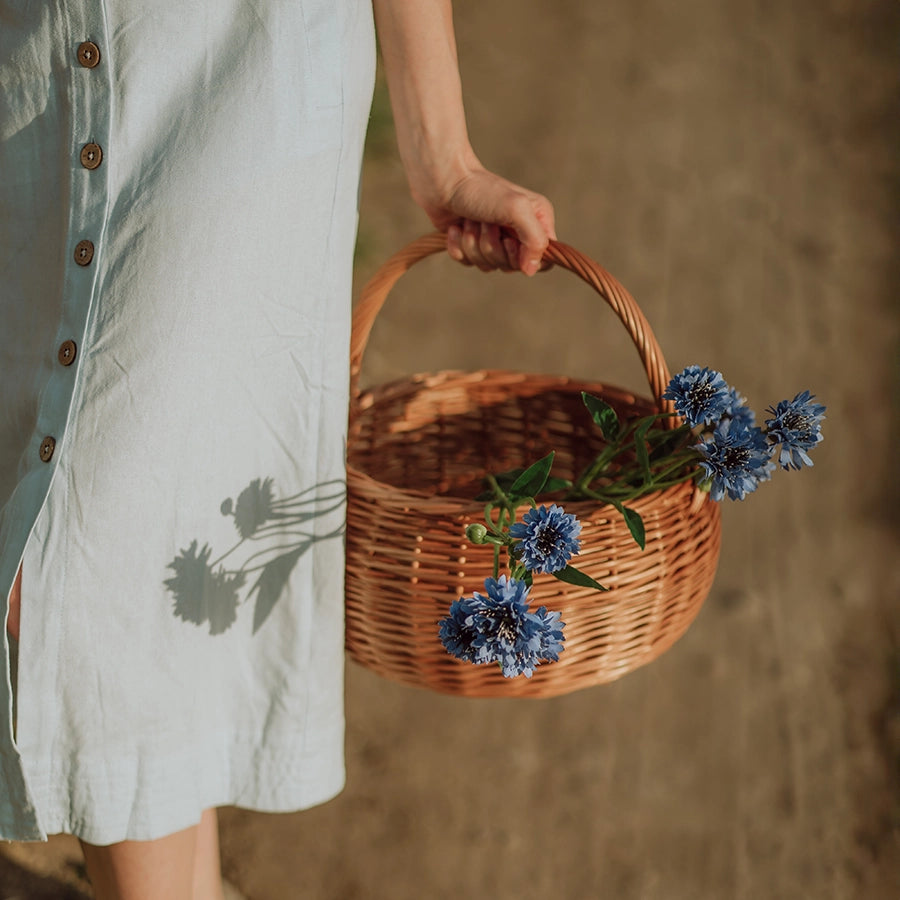 Kobieta w naturalnym otoczniku letniego pola trzyma ręcznie pleciony kosz z wikliny - produkt z oferty sklepu MUNIA naturalnie, specjalizującego się w ekologicznych wyrobach wiklinowych.