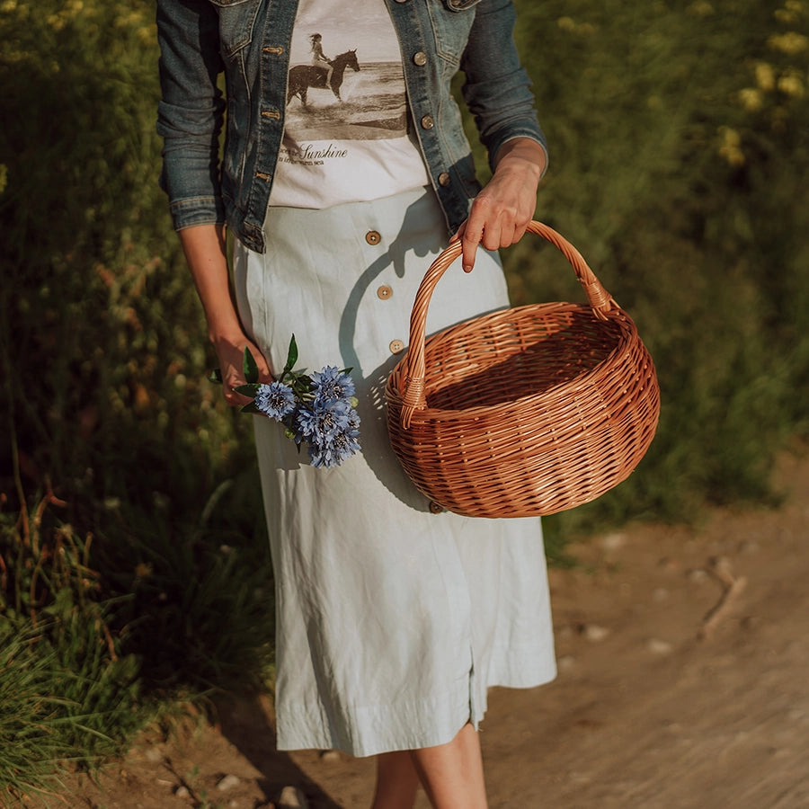 Kobieta w naturalnym otoczniku letniego pola trzyma ręcznie pleciony kosz z wikliny - produkt z oferty sklepu MUNIA naturalnie, specjalizującego się w ekologicznych wyrobach wiklinowych.
