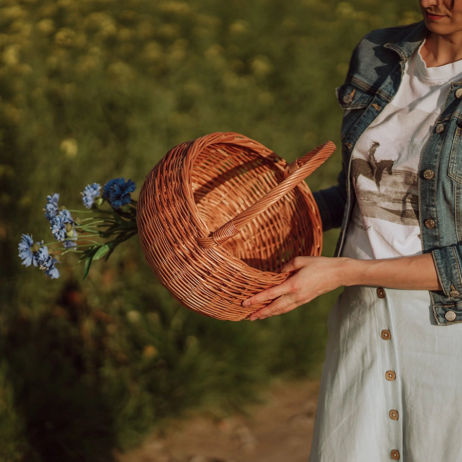 Kobieta w naturalnym otoczniku letniego pola trzyma ręcznie pleciony kosz z wikliny - produkt z oferty sklepu MUNIA naturalnie, specjalizującego się w ekologicznych wyrobach wiklinowych.