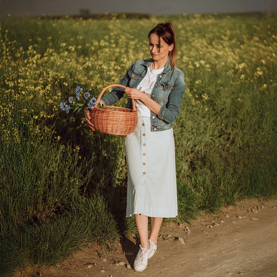 Kobieta w naturalnym otoczniku letniego pola trzyma ręcznie pleciony kosz z wikliny - produkt z oferty sklepu MUNIA naturalnie, specjalizującego się w ekologicznych wyrobach wiklinowych.