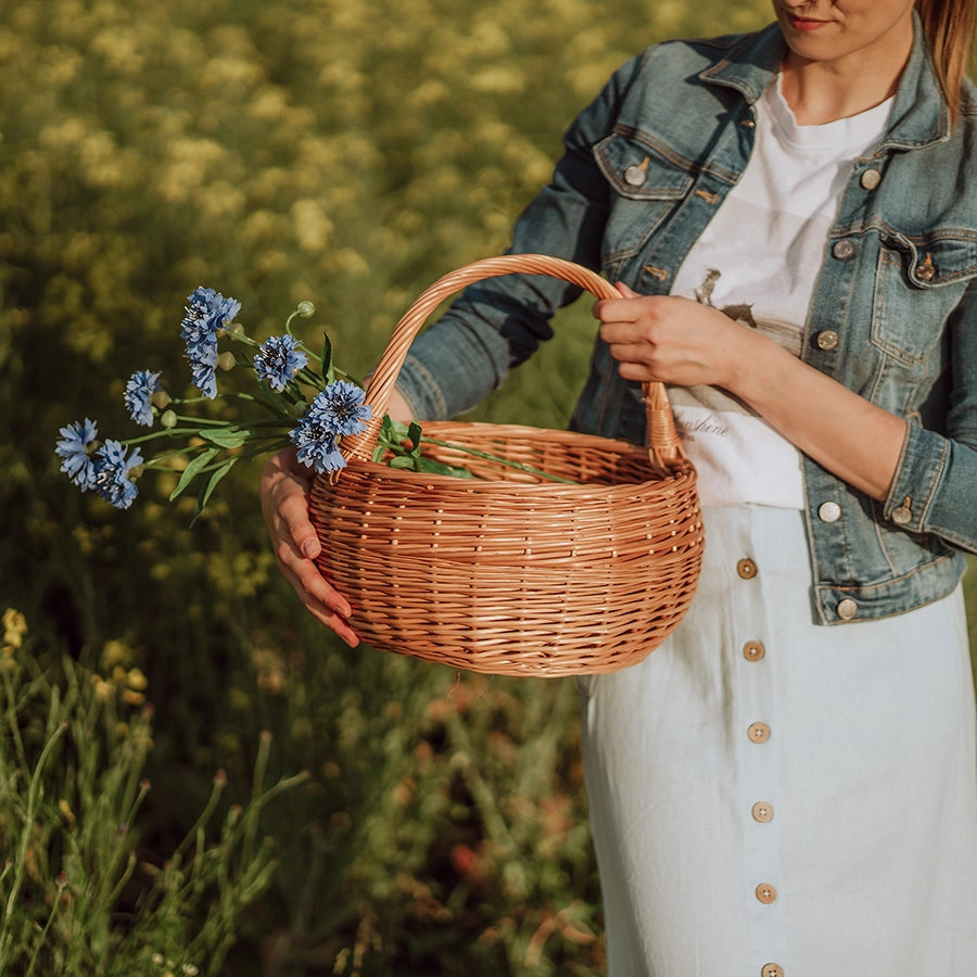 Kobieta w naturalnym otoczniku letniego pola trzyma ręcznie pleciony kosz z wikliny - produkt z oferty sklepu MUNIA naturalnie, specjalizującego się w ekologicznych wyrobach wiklinowych.