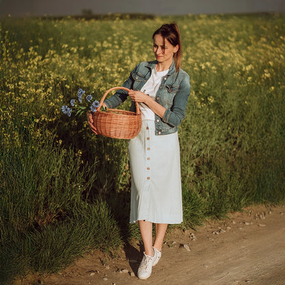 Kobieta w naturalnym otoczniku letniego pola trzyma ręcznie pleciony kosz z wikliny - produkt z oferty sklepu MUNIA naturalnie, specjalizującego się w ekologicznych wyrobach wiklinowych.